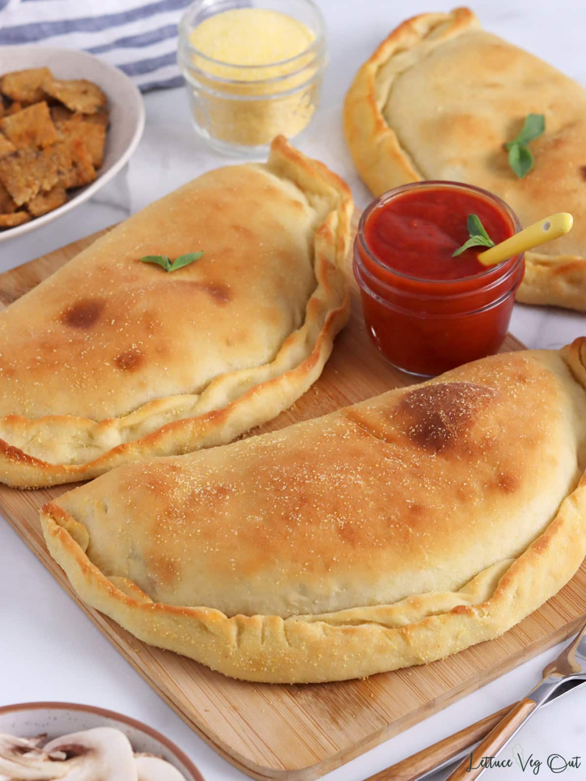 Two baked calzones on a wood board with dishes of ingredients around (tempeh, mushrooms, cornmeal, pizza sauce).