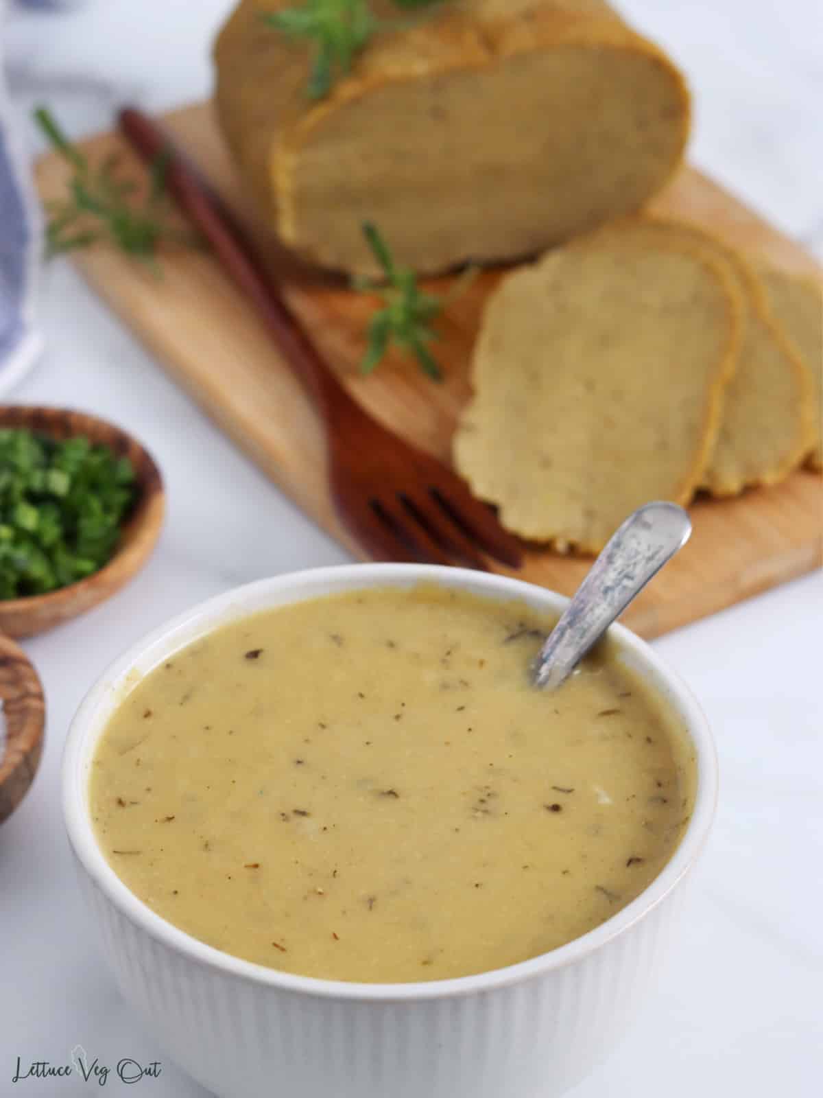 Bowl of gravy with spoon in it, in front of a seitan turkey on wood board.
