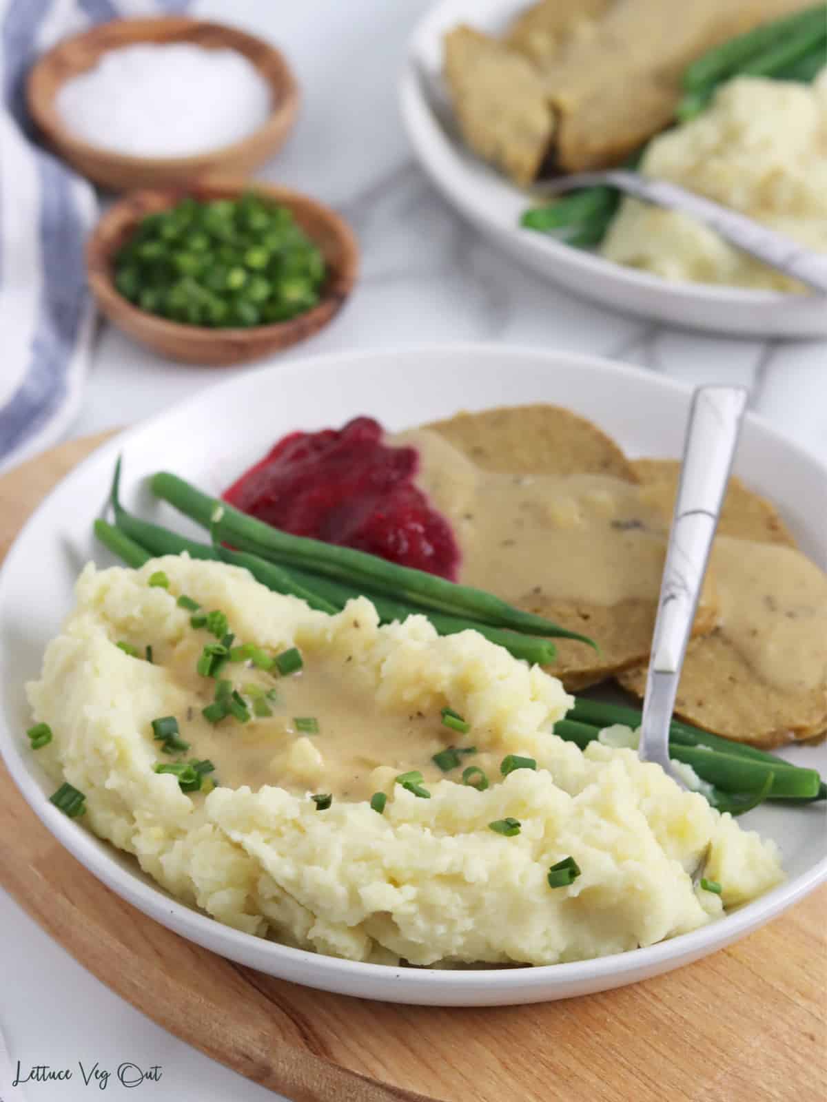 Plate with pile of fluffy, creamy mashed potatoes topped with gravy and chives with some vegan turkey, green beans and cranberry sauce on back of plate.