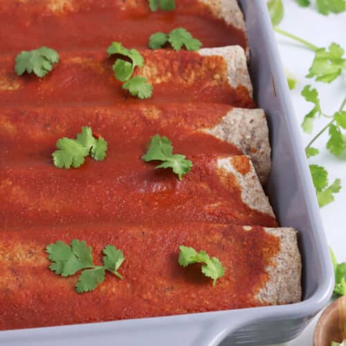 Close up of a baking dish filled with vegan enchiladas topped with red sauce and cilantro garnish.