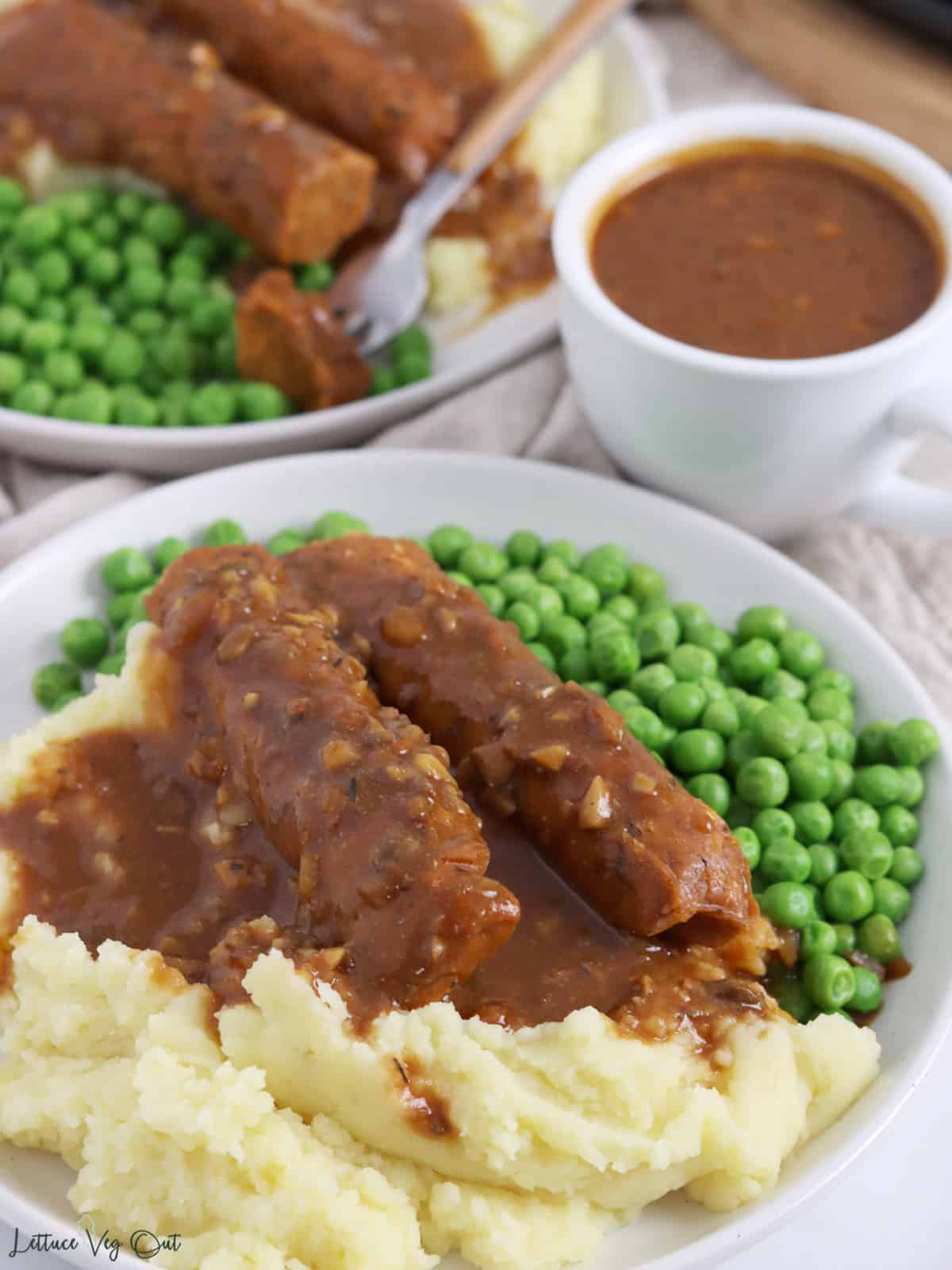 Plate of sausage, mashed potatoes, peas and gravy with small dish of gravy in background.