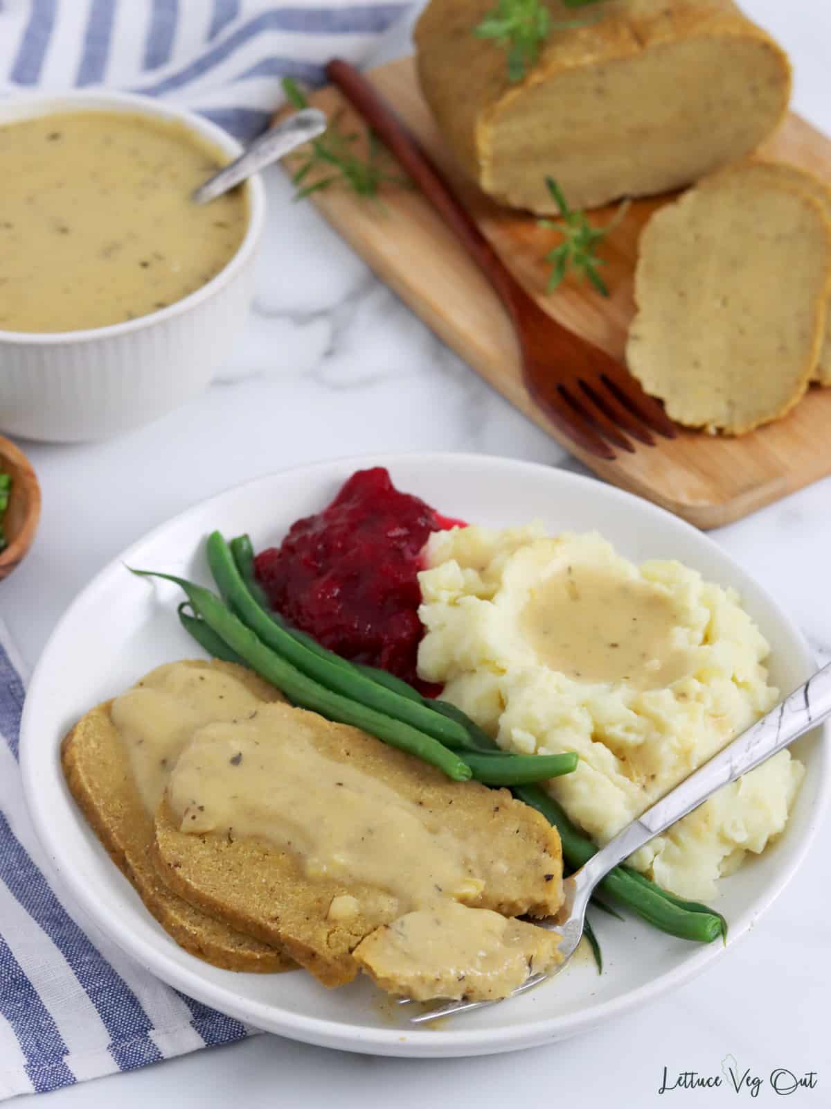 Plate of vegan turkey roast with gravy, potatoes, green beans and cranberry sauce with serving board of roast in background.