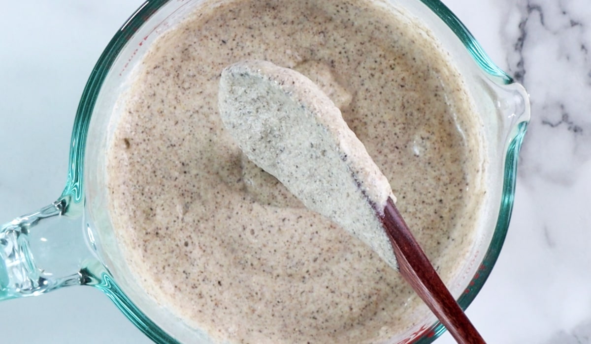 Spoonful of thick buckwheat pancake batter held up over a glass measuring cup of batter.