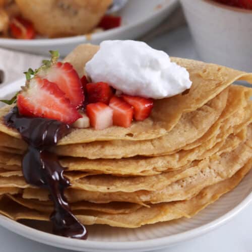 Close up of a stack of folded red lentil pancakes topped with strawberries, whipped cream and chocolate drizzle.
