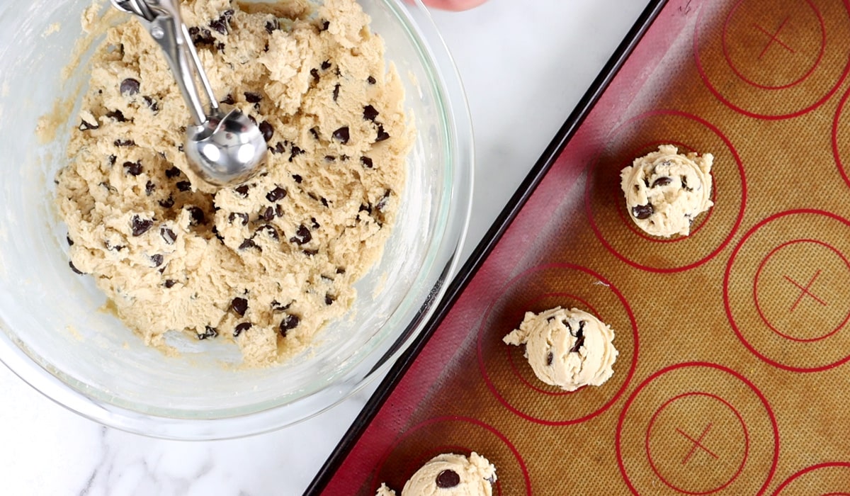 Bowl of chocolate chip cookie dough with a hand scooping dough and a baking tray with three cookie dough balls on it.