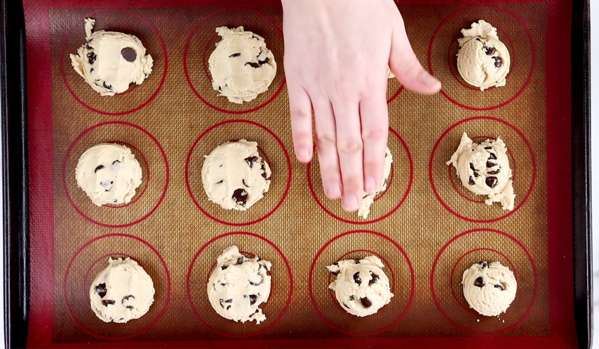 Baking tray with raw chocolate chip cookies and a hand pressing down on a ball of cookie dough.