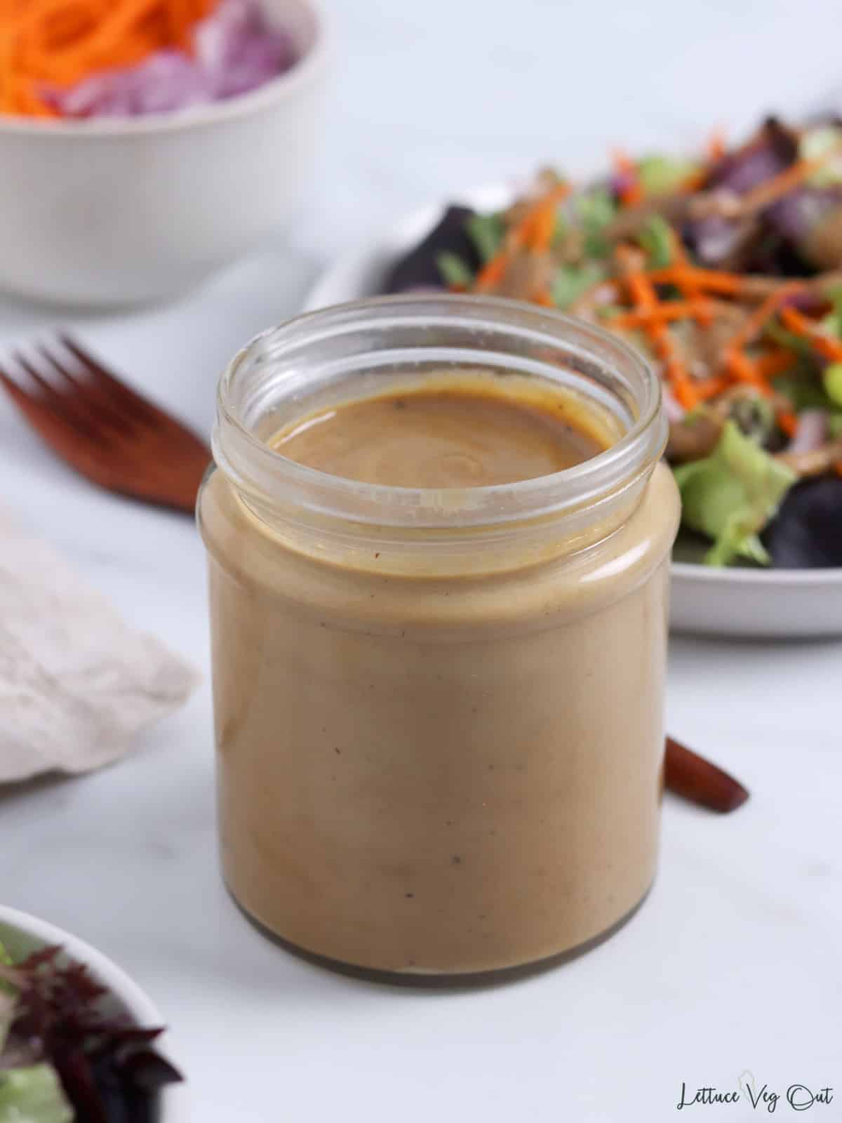 Jar of tahini balsamic dressing with blurry plates of salad surrounding it.