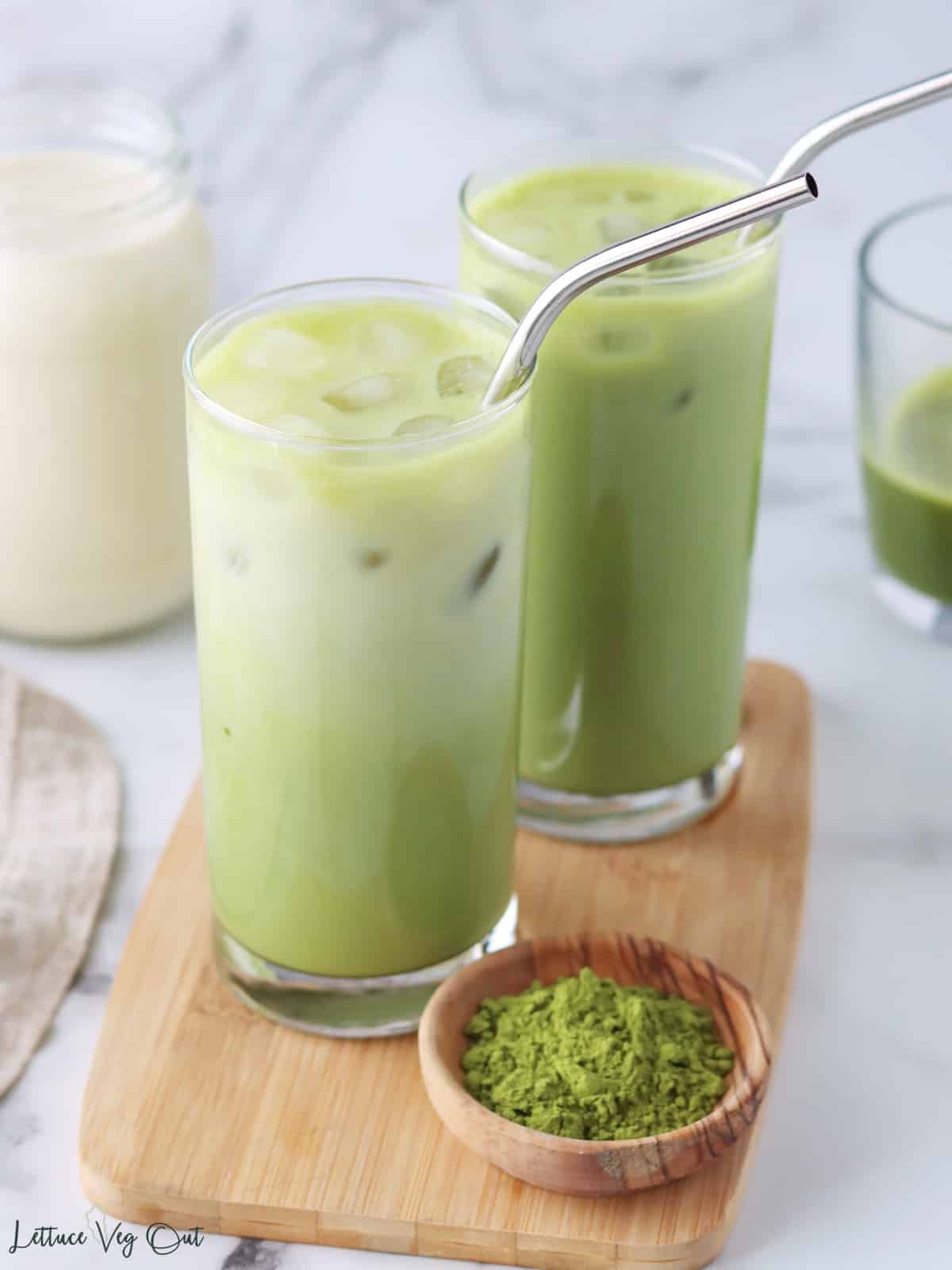 Two tall glasses of iced matcha latte on wood board with small bowl of matcha powder.