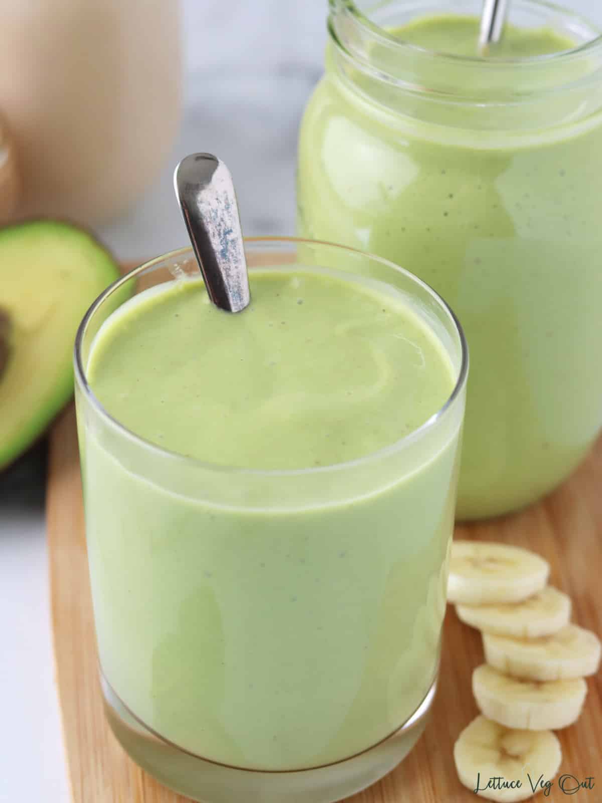 Creamy green smoothie in a glass with a spoon and second glass behind with banana slices garnishing.