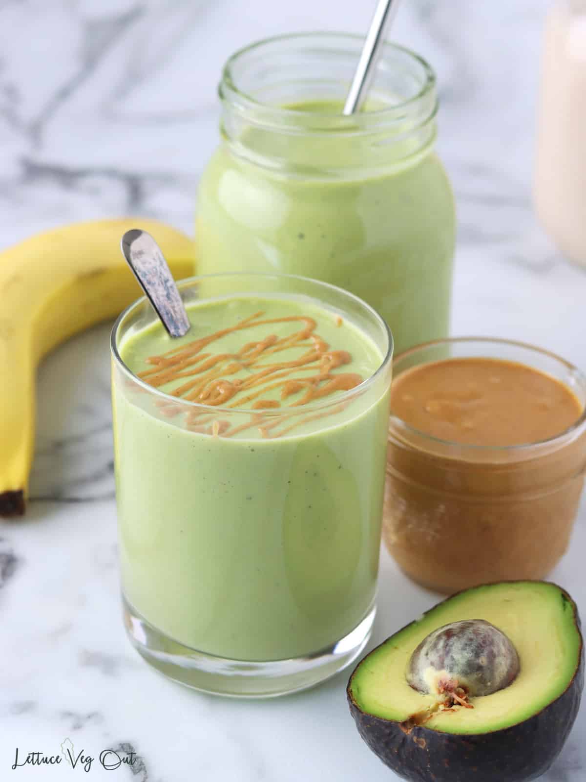 Two glass of green smoothie with metal straws and peanut butter drizzle on one smoothie with banana, peanut butter and avocado decorating around glasses.