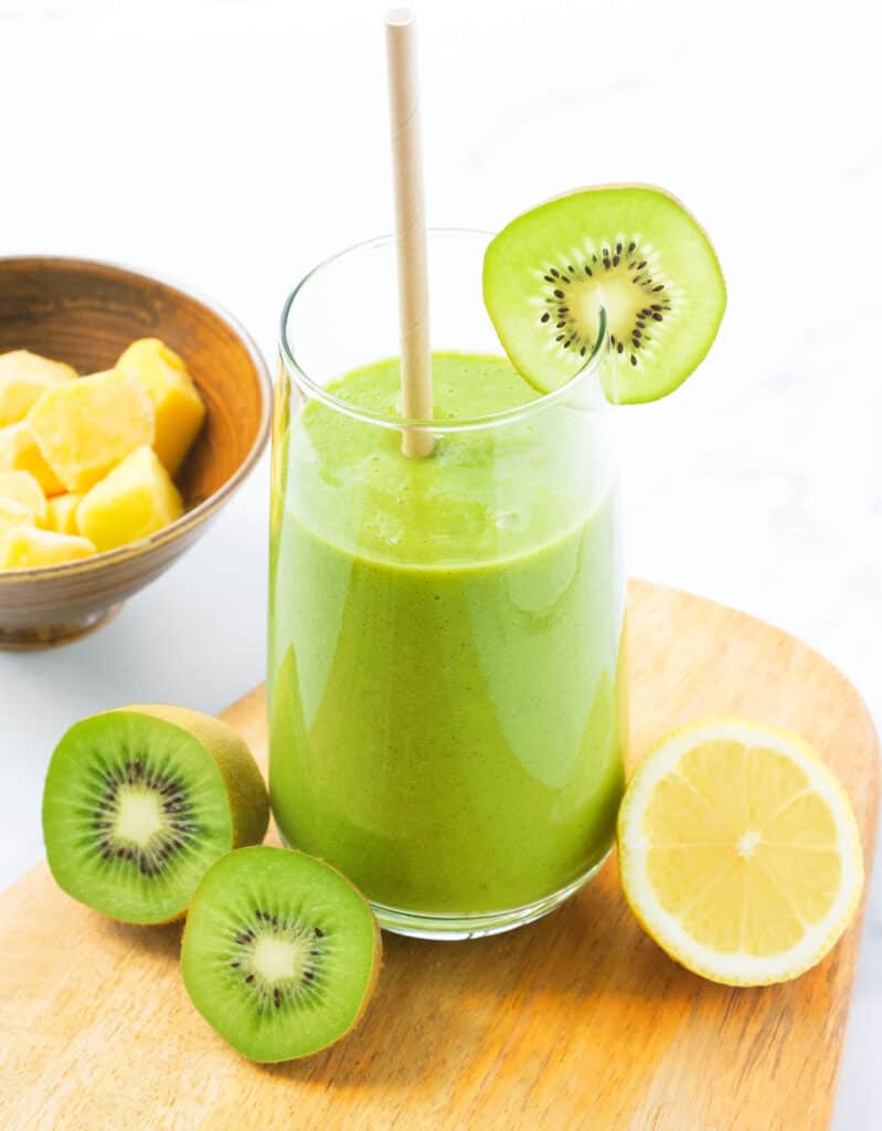 Glass of green smoothie with a paper straw and decorated with kiwi slices and lemon.