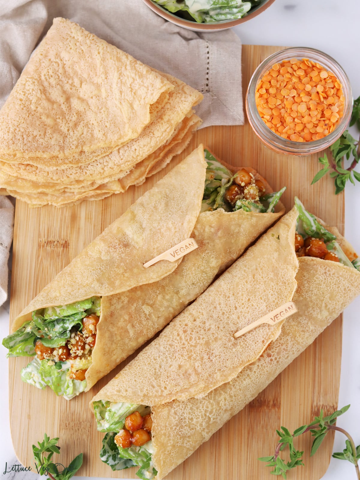 Two red lentil tortillas wrapped with salad on a wood board next to a folded stack of more lentil tortillas.