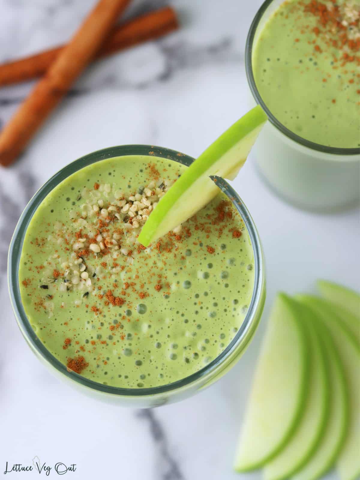 Close up of the top of a glass filled with creamy green smoothie and topped with cinnamon and hemp seeds with sliced apple garnish.