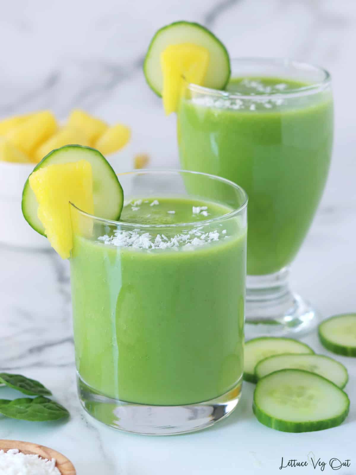 Two glasses of pineapple green smoothie with cucumber slices and a bowl of pineapple decorating around glasses.