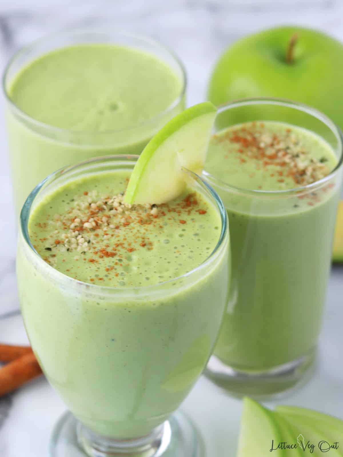 Three glasses with green apple smoothie with two garnished with apple, cinnamon and hemp seeds.