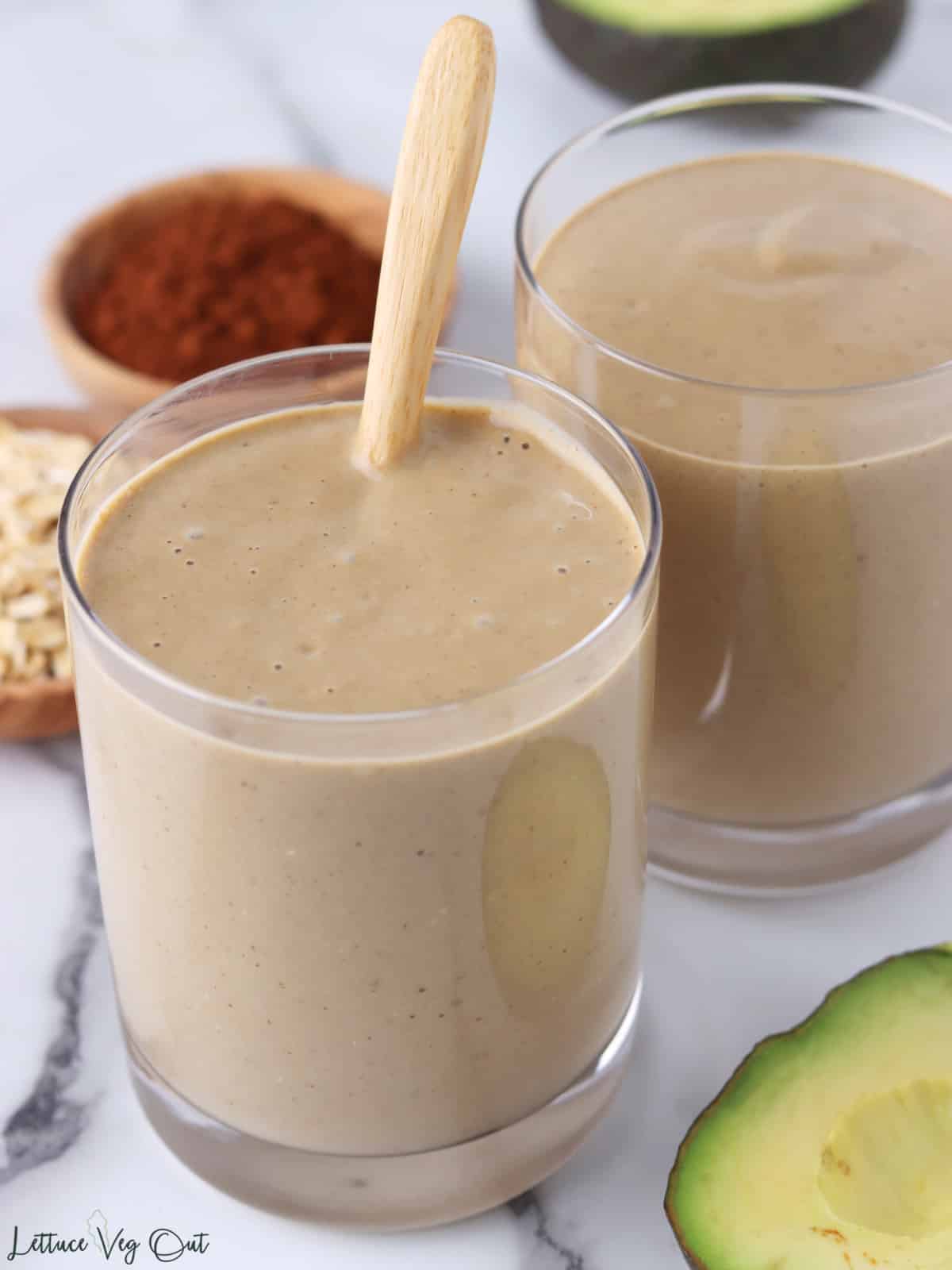 Two glasses of chocolate avocado oatmeal smoothie with avocado and small dish of cocoa powder around the glasses.