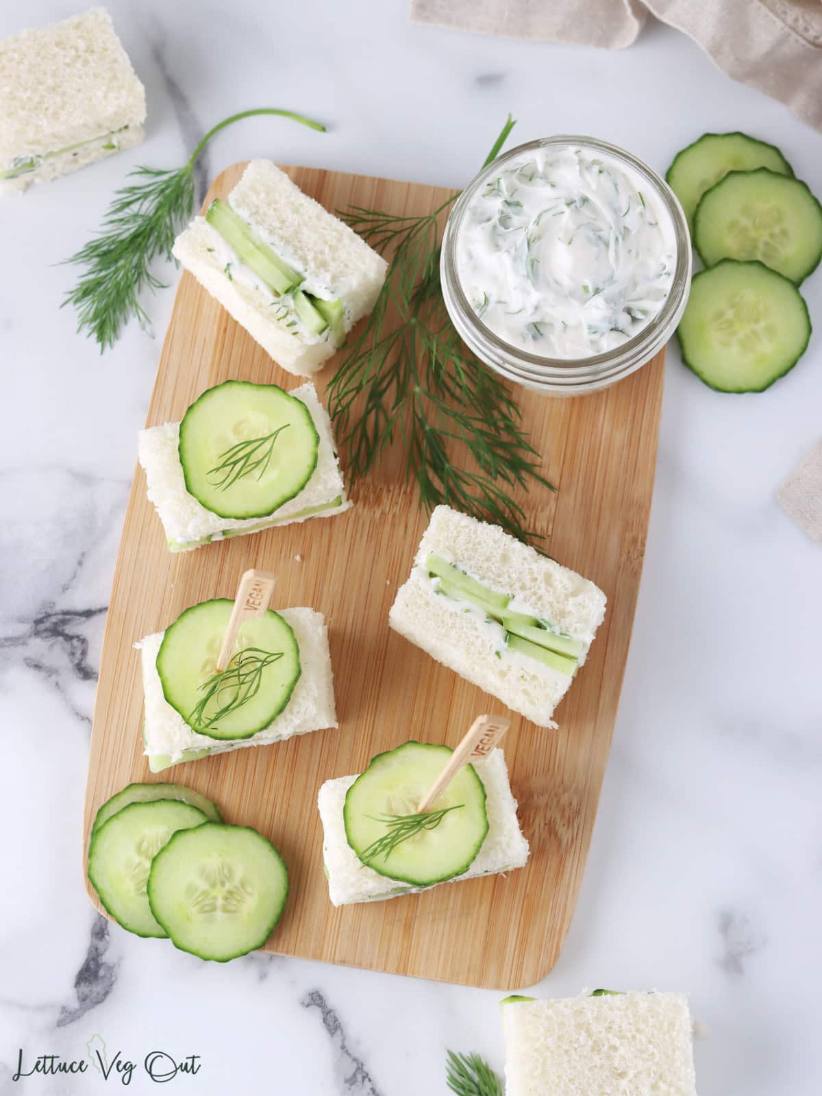 Small cucumber tea sandwiches arranged on a wood board with cucumber and dill garnish plus small jar of cream cheese.