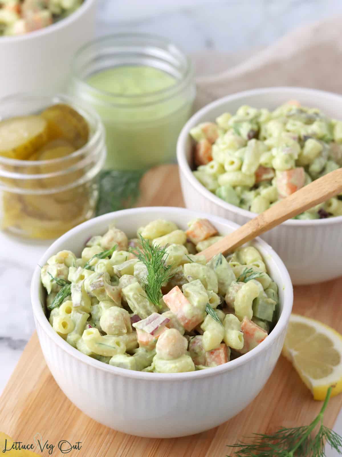 Two small bowls of dill pickle macaroni salad on wood board with lemon and pickle garnishes.