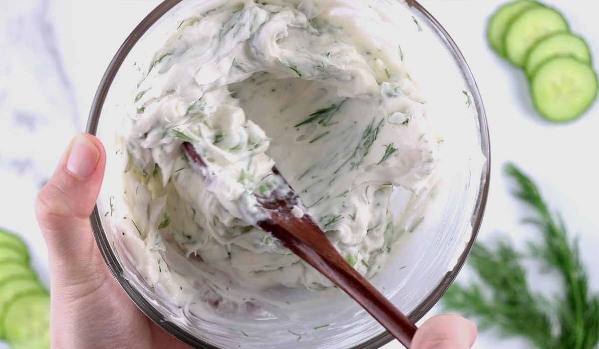 Hands stirring herb cream cheese in a glass bowl with a wood spoon.