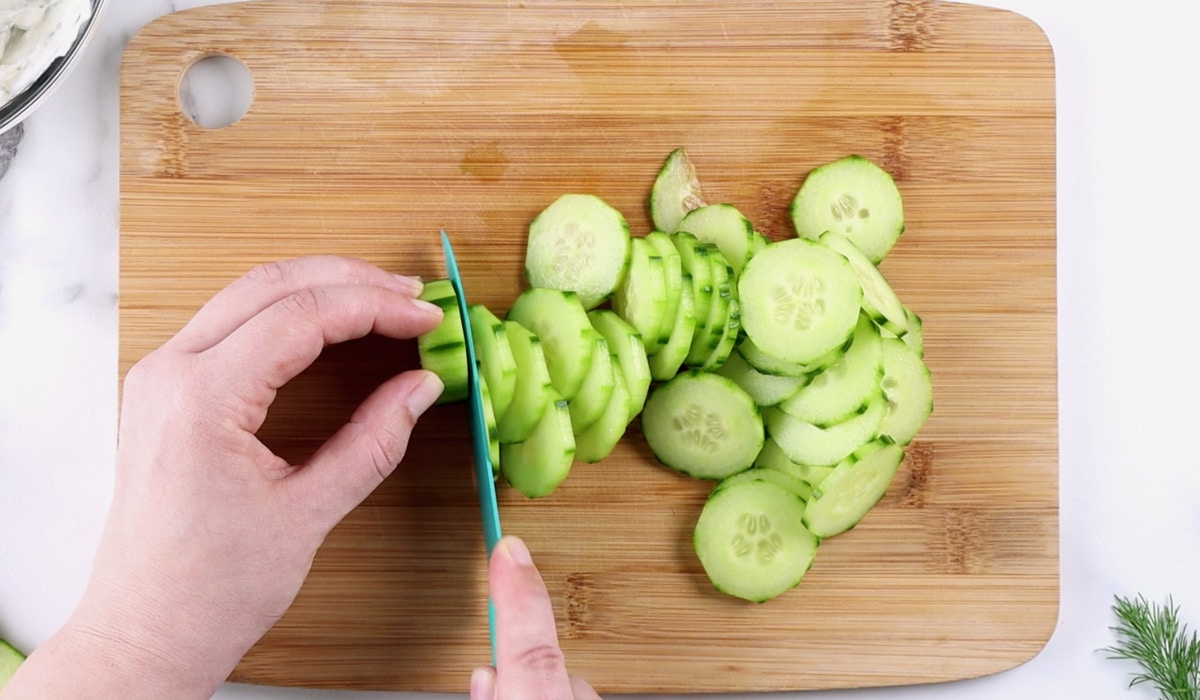 Hands slicing cucumber on a wood board.