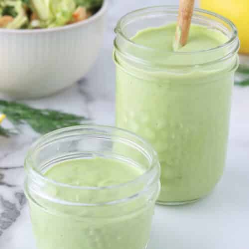 Close up of two jars filled with creamy, green tahini dill dressing with a wood spoon in one jar.
