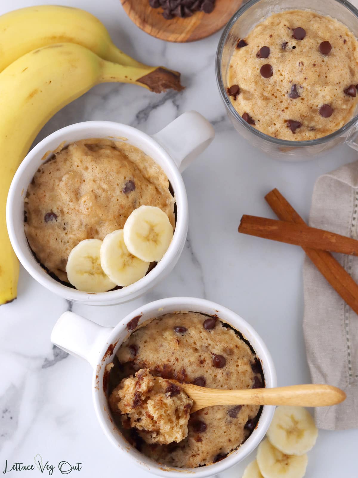 Three mugs with banana mug cake made with mini chocolate chips, one mug has spoonful of cake resting on it and another is garnished with banana slices.