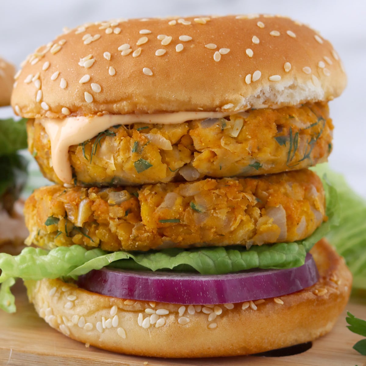 Close up of an assembled chickpea burger with two patties, a fluffy bun, sriracha mayo, red onion and lettuce.
