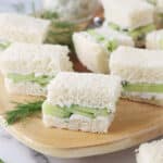 Close up of small cucumber cream cheese tea sandwiches on wood board with dill garnish.