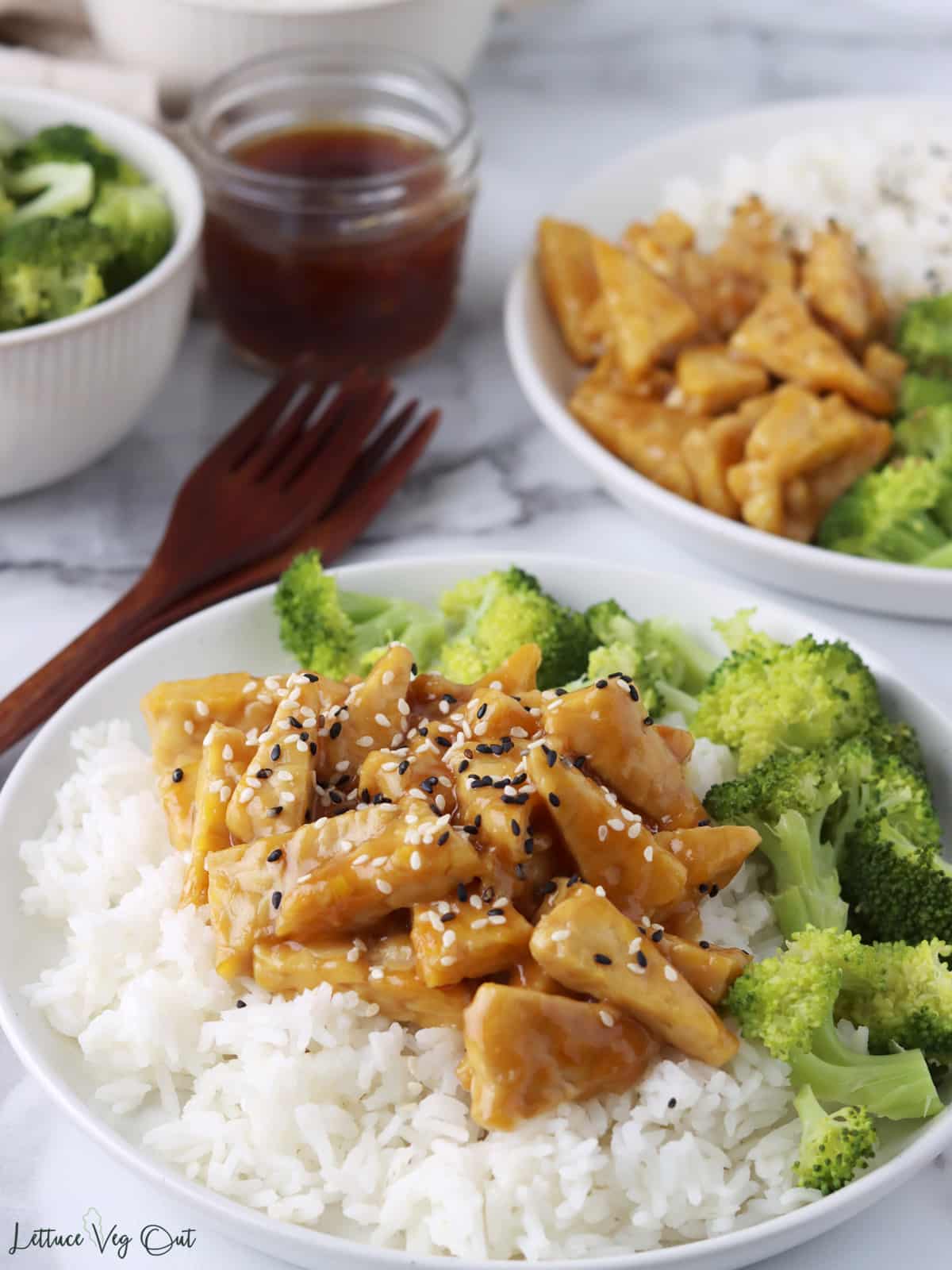 Plate of teriyaki tempeh over rice with broccoli and second plate blurred in back.