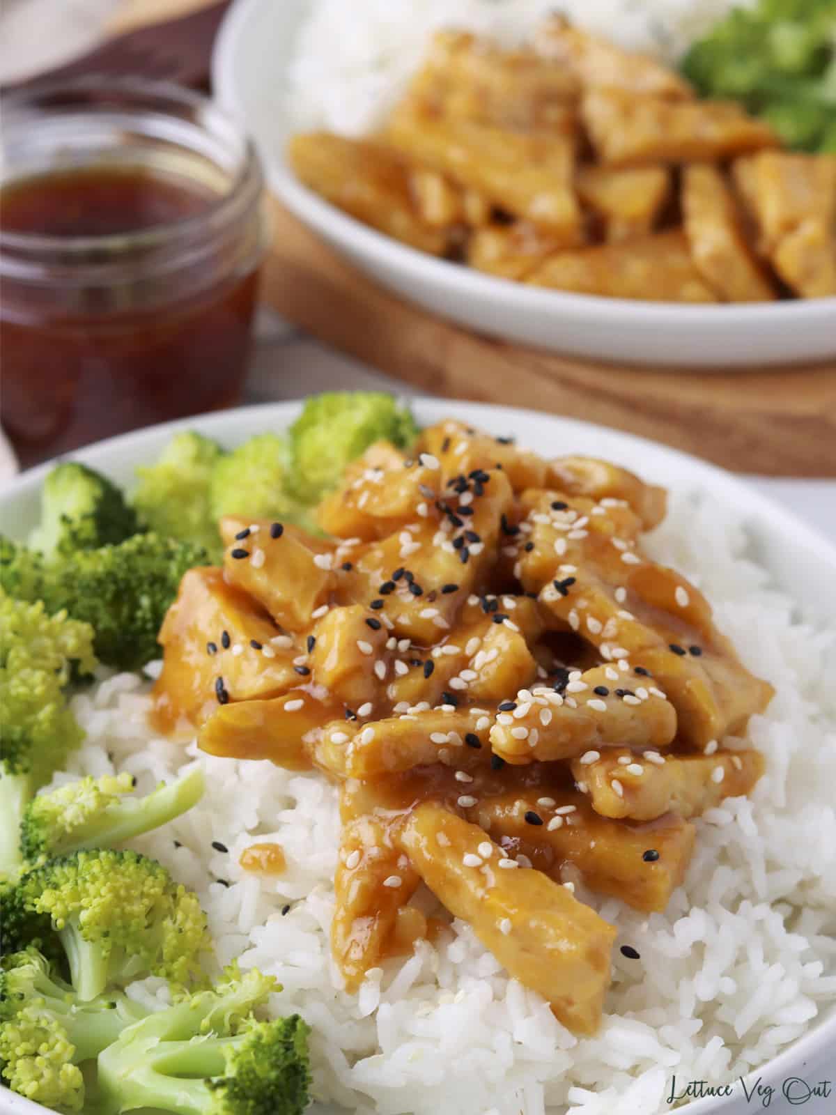 Teriyaki tempeh, rice and broccoli on a plate with second plate and jar of teriyaki sauce in back.