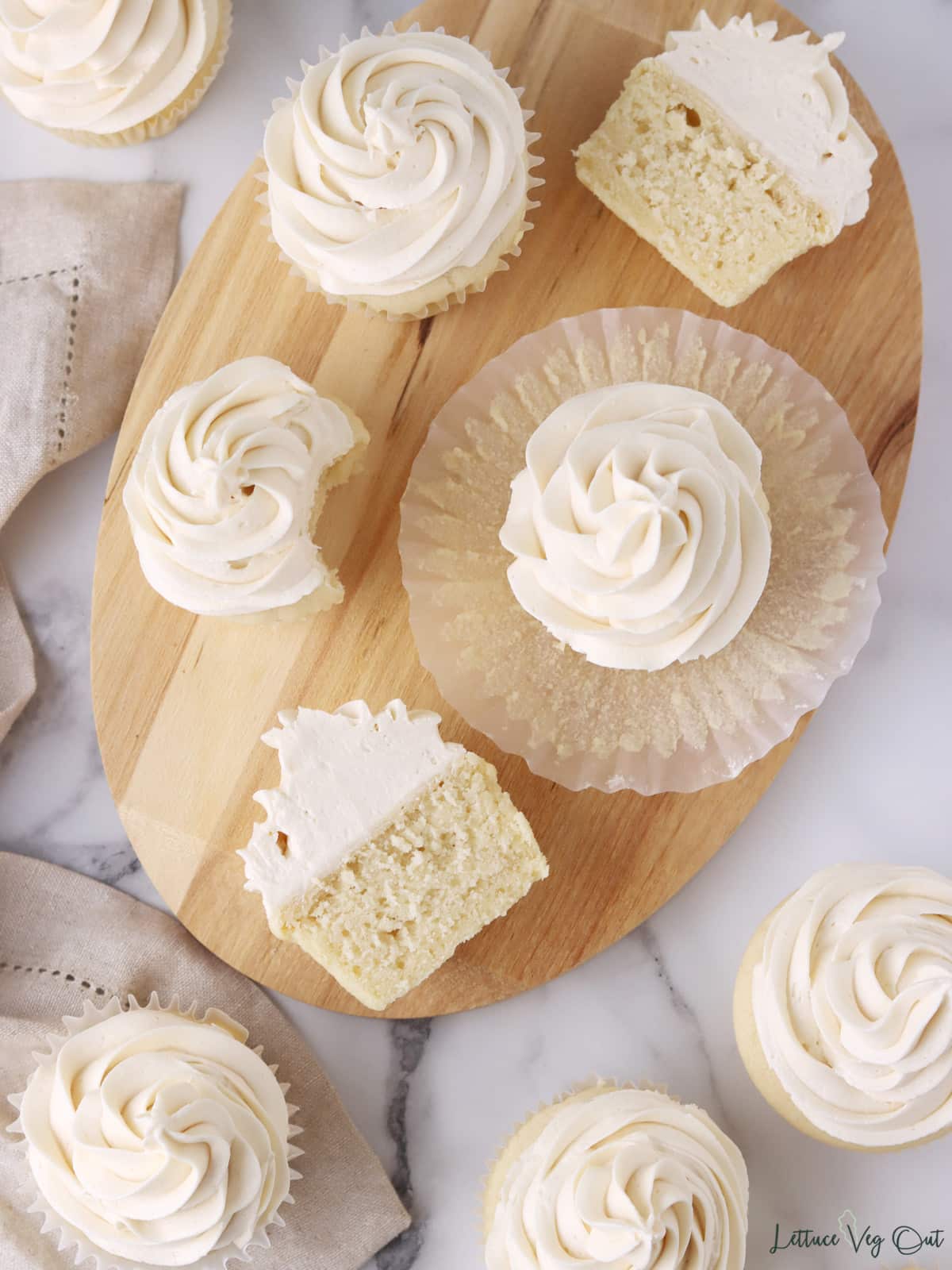 Whole and halved vanilla cupcakes on a wood board, some cupcakes on their side, all topped with a large swirl of vanilla buttercream frosting.