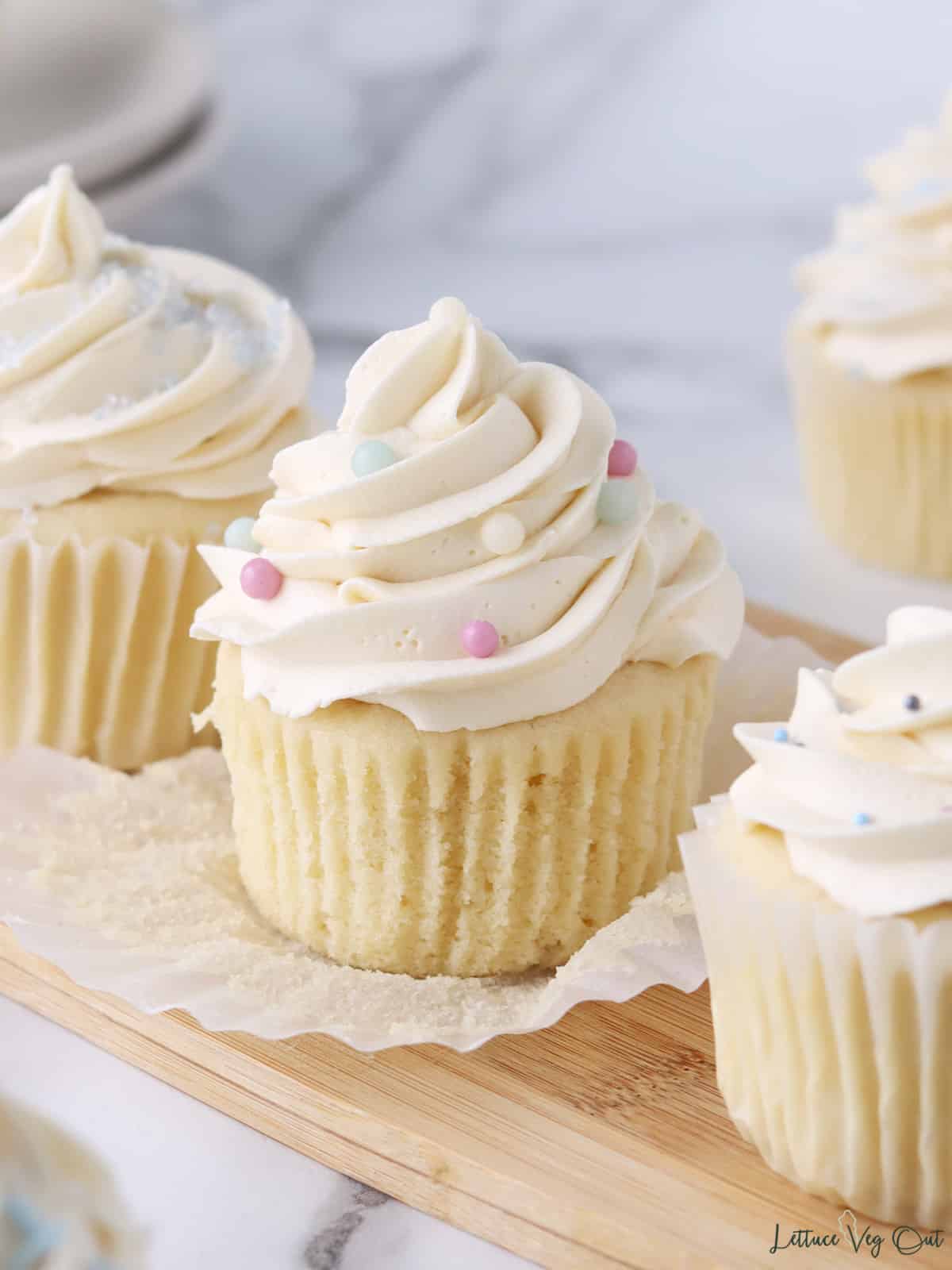 Close up of a vanilla cupcake topped with vanilla buttercream and large pale sprinkles, surrounded by other cupcakes.