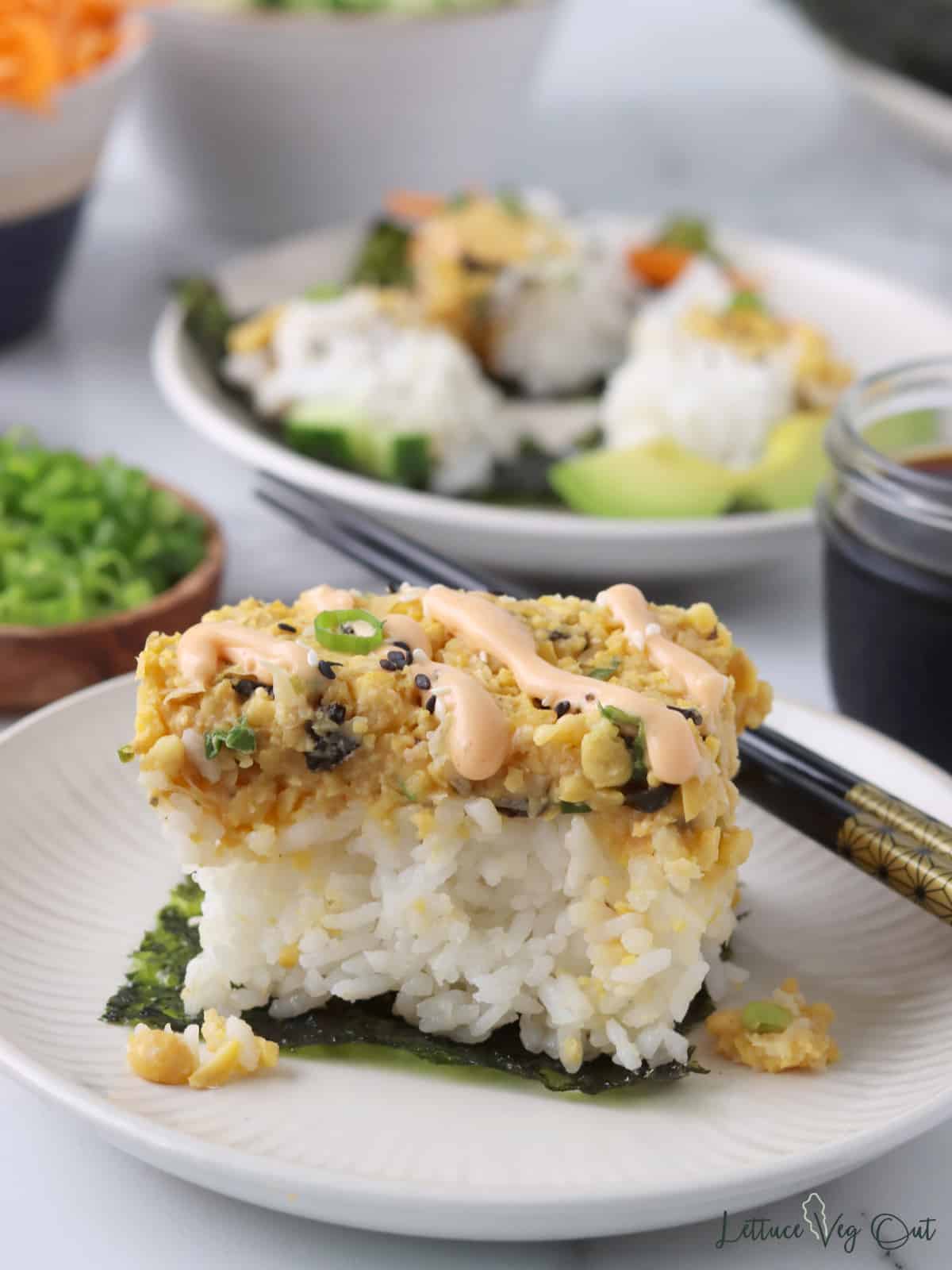 Piece of sushi bake (cooked rice topped with mashed chickpea "tuna") on a square of nori with chopsticks.