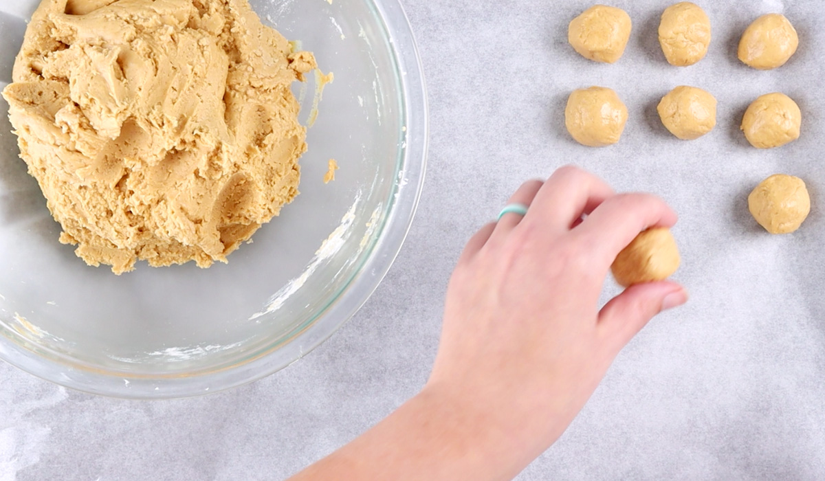 Hand placing a ball of rolled peanut butter filling onto a baking tray, next to a bowl of peanut butter ball dough.