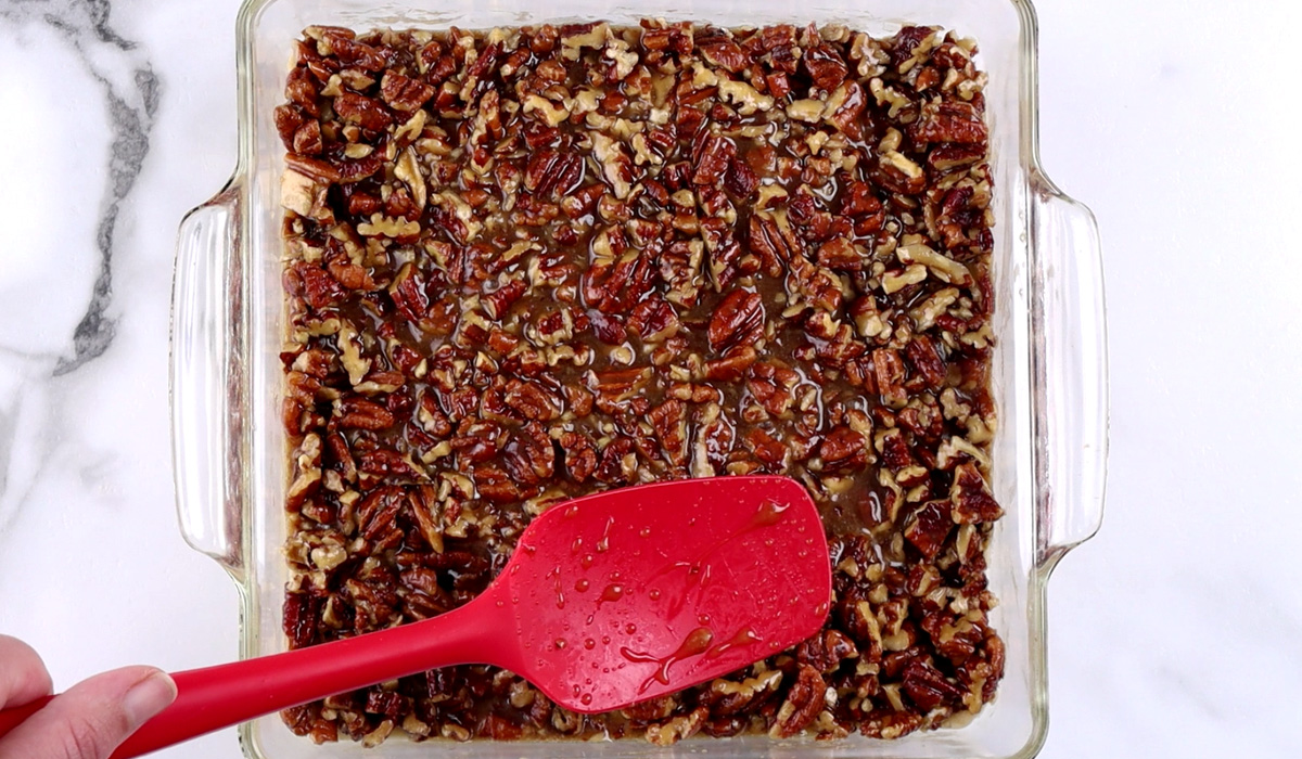 Red spatula spreading pecan pie filling over partially baked crust in square, glass baking dish.