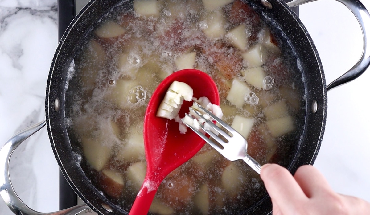 Fork mashing a cooked piece of potato on a red spoon over a pot of cooked potatoes.